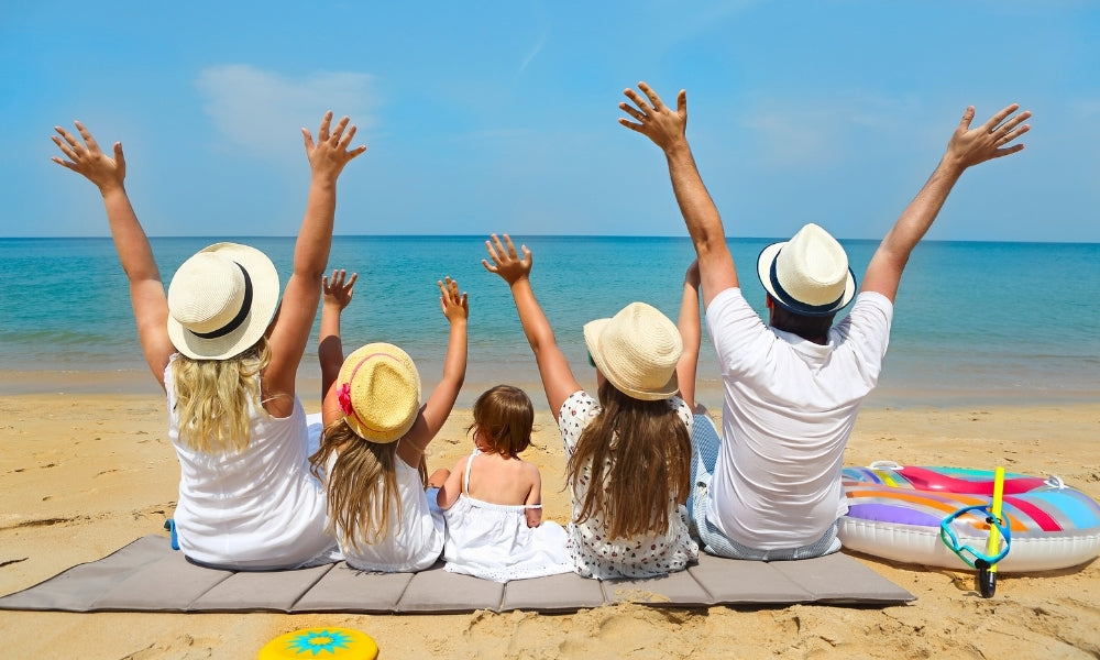 Familia disfrutando las vacaciones de verano en la playa, sentados frente al mar con los brazos en alto, representando descanso, tiempo en familia y calidad de vida durante el periodo de vacaciones.