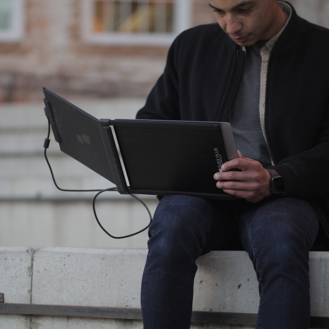 Estudiante al aire libre utilizando monitor portátil conectado a notebook para estudiar en plaza