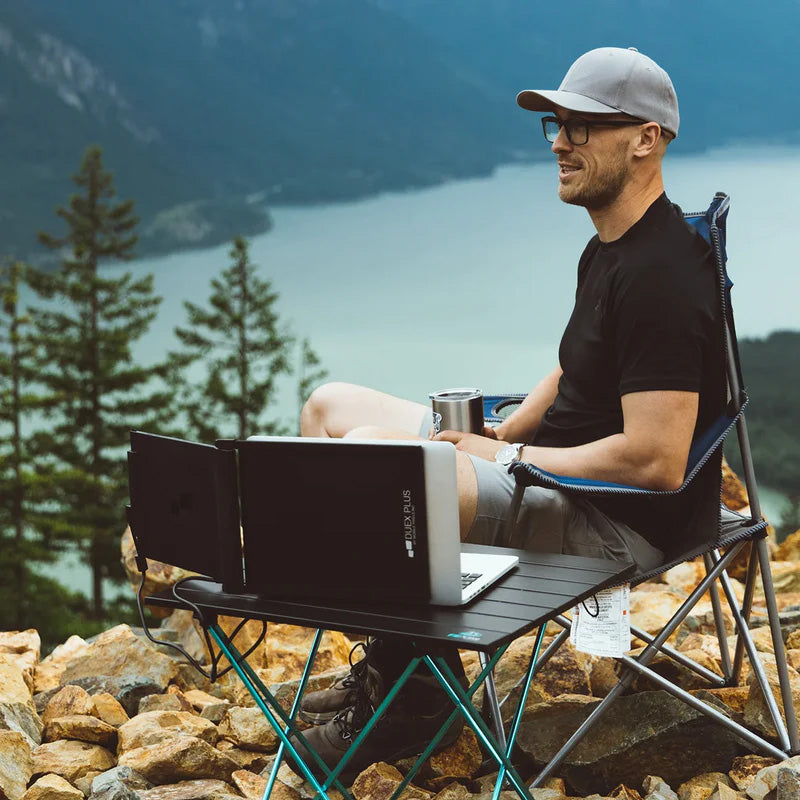 Persona acampando y trabajando al aire libre con monitor portátil Mobile Pixels Duex Plus conectado a laptop; setup ideal para trabajo remoto, estilo de vida nómada digital y productividad en entornos naturales.