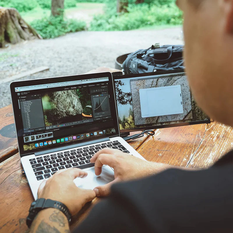 Persona trabajando cómodamente al aire libre en el bosque con monitor portátil Mobile Pixels Duex Plus conectado a laptop; edición de fotografías en pantalla extendida ideal para fotógrafos, creadores visuales y nómadas digitales.