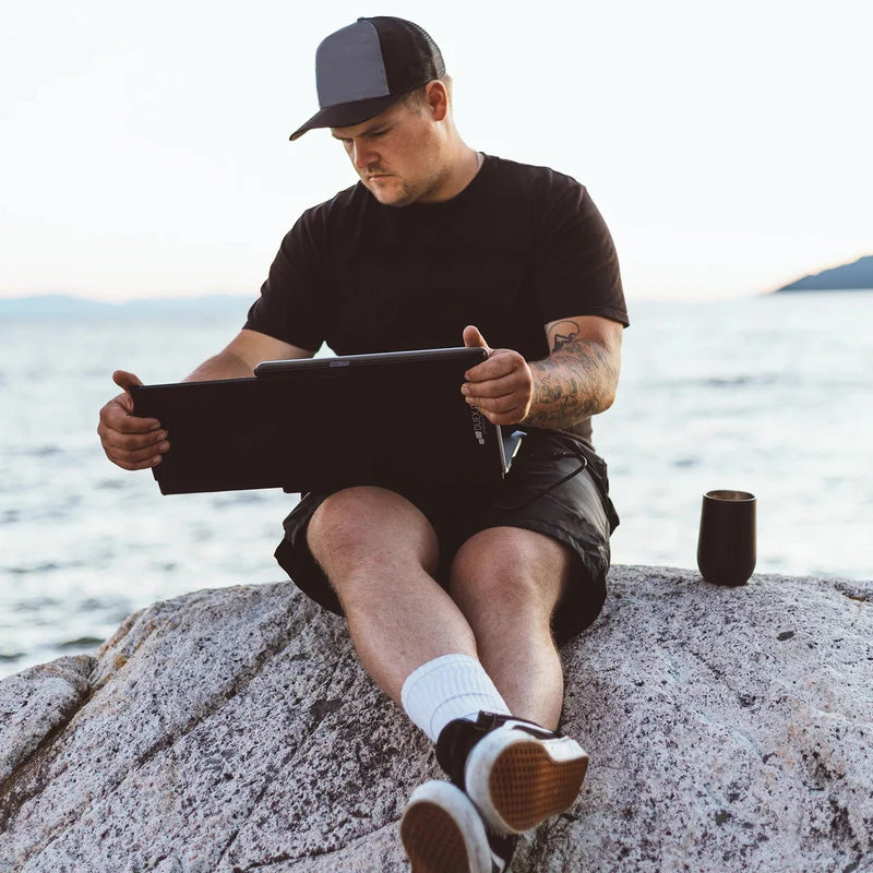 Persona trabajando al aire libre desde la orilla de un lago con monitor portátil Mobile Pixels Duex Plus conectado a laptop; pantalla extendida ideal para trabajo remoto, estilo de vida nómada digital y productividad en entornos naturales.
