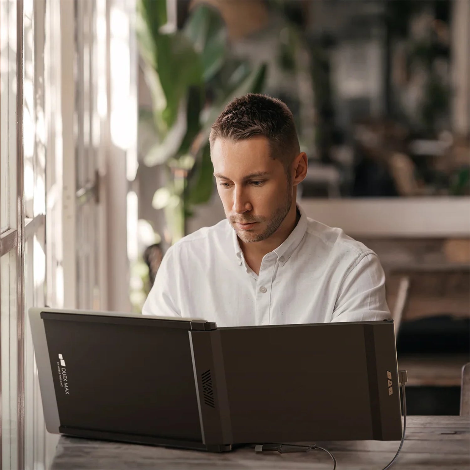 Joven trabajando en un espacio de coworking con monitor portátil Mobile Pixels Duex Max conectado a su laptop; pantalla extendida ideal para productividad profesional, multitareas y trabajo remoto en entornos colaborativos.