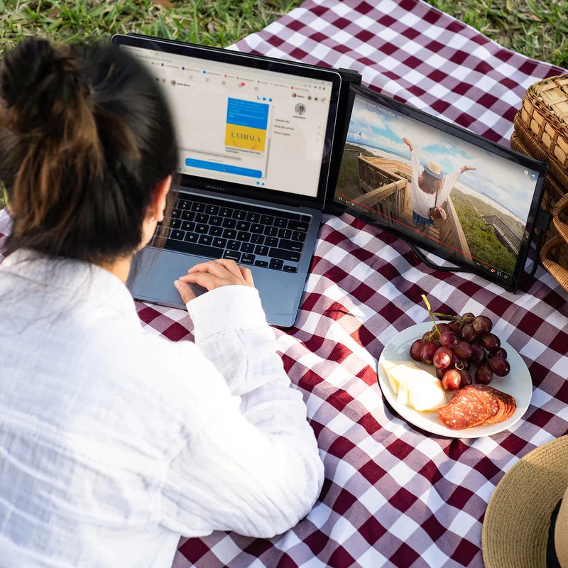 Joven trabajando al aire libre durante un picnic con monitor portátil Mobile Pixels Duex Max; pantalla extendida ideal para combinar productividad y entretenimiento, viendo videos favoritos mientras trabaja desde su laptop.